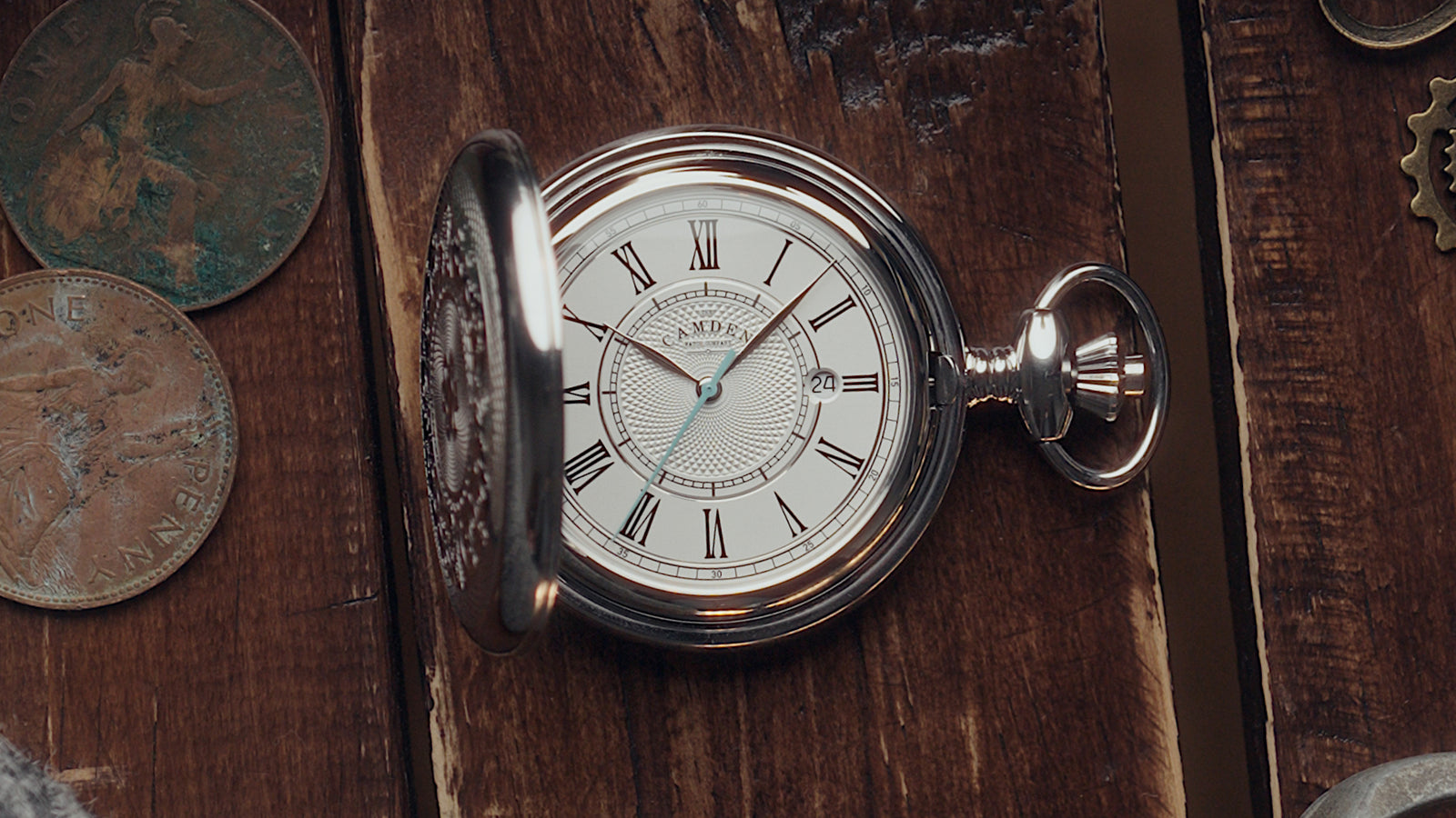 The Camden watch company steel pocket watch with a white dial and black Roman numerals, shown open on a rustic dark wooden surface with old coins nearby.