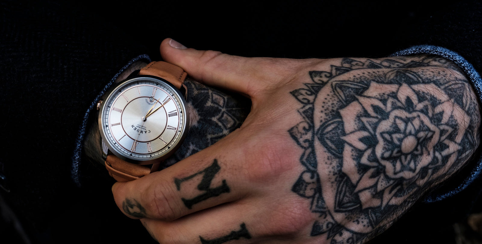 Hand with tattoos holding a wristwatch against a dark background